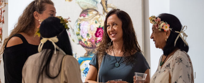 A group of four women stand together at an art gallery, chatting in front of a large artwork featuring ornate, floral-patterned teapots stacked in a whimsical arrangement. Two of the women wear floral headbands, one holds a drink, and another carries a handbag. The setting feels lively and social, with warm lighting and a festive atmosphere.