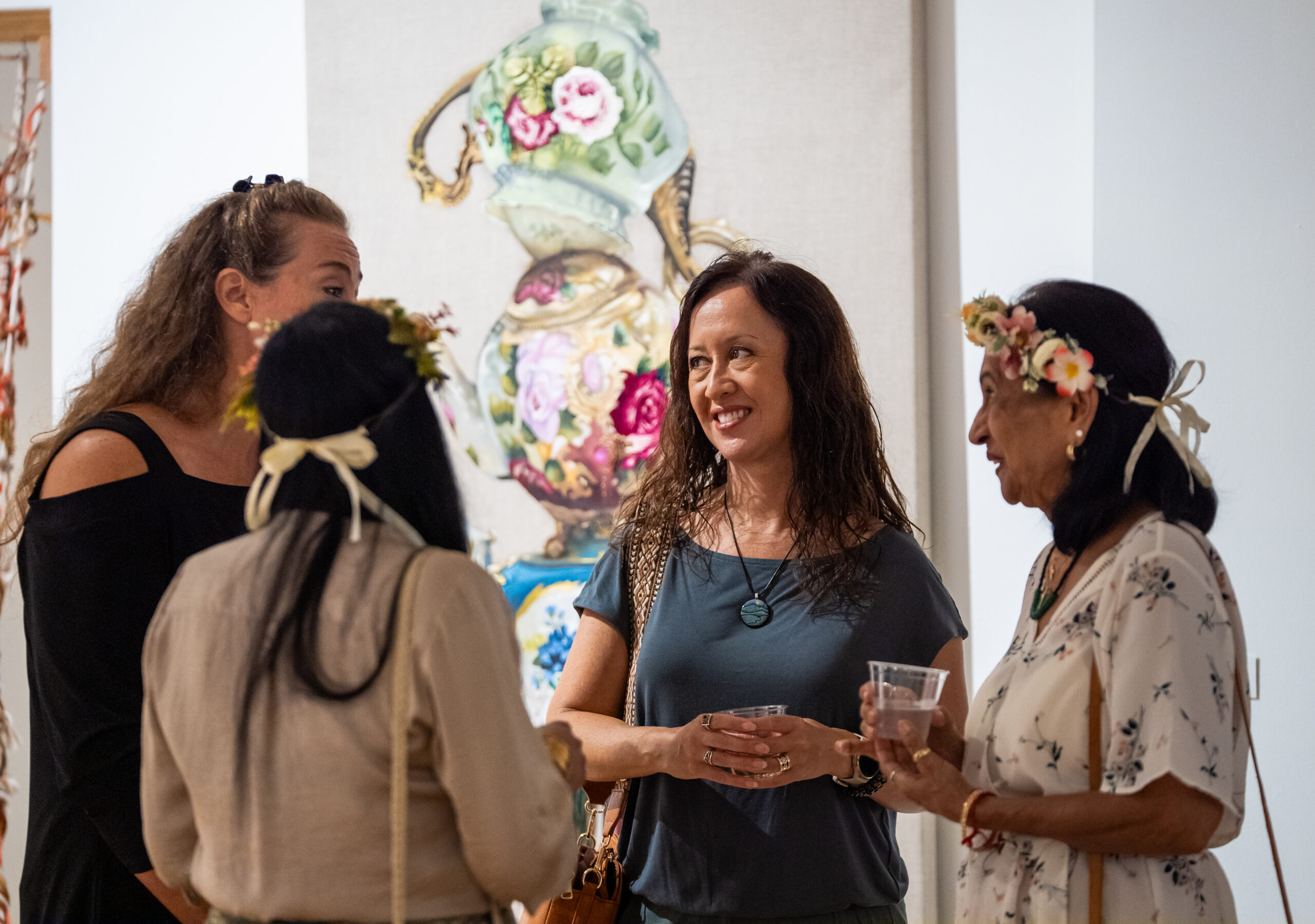A group of four women stand together at an art gallery, chatting in front of a large artwork featuring ornate, floral-patterned teapots stacked in a whimsical arrangement. Two of the women wear floral headbands, one holds a drink, and another carries a handbag. The setting feels lively and social, with warm lighting and a festive atmosphere.
