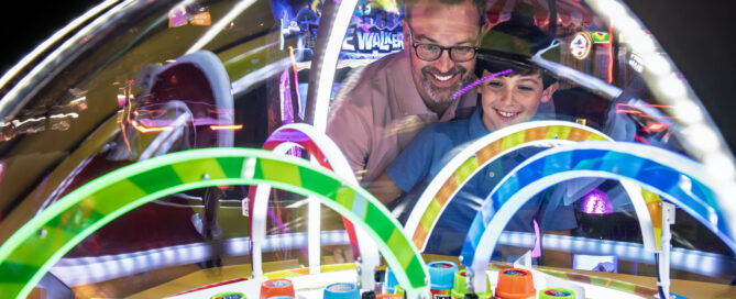 Two people stand close together at a brightly lit arcade machine with a transparent dome filled with colorful rings and stacks of “150 TICKETS” cylinders. Neon lights and reflections create a lively, energetic arcade atmosphere as they play the game.