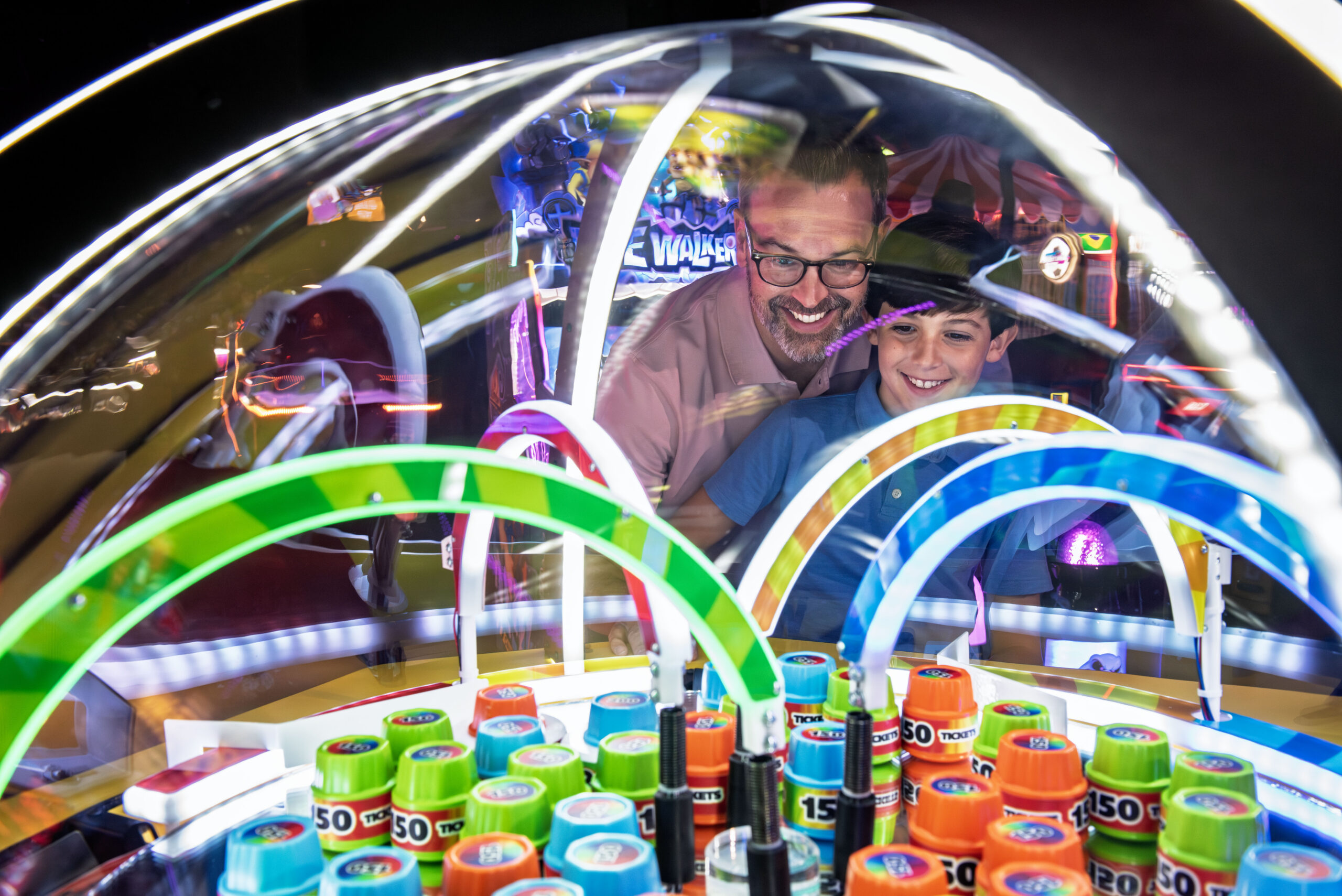 Two people stand close together at a brightly lit arcade machine with a transparent dome filled with colorful rings and stacks of “150 TICKETS” cylinders. Neon lights and reflections create a lively, energetic arcade atmosphere as they play the game.